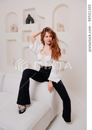 an attractive woman in a white top and black pants sits on a white sofa. an attractive woman in a white top and black pants sits on a white sofa. 99501305