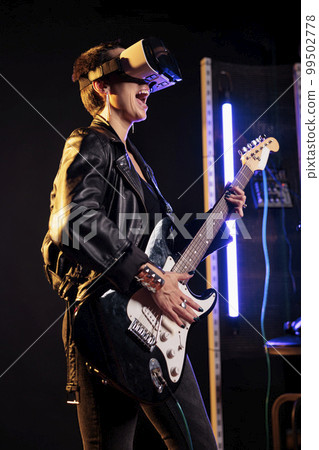 Rebel performer playing heavy metal song using electric guitar while wearing vr headset to have rock concert simulation, enjoying experience in music studio. Woman musician working at new grunge album 99502778