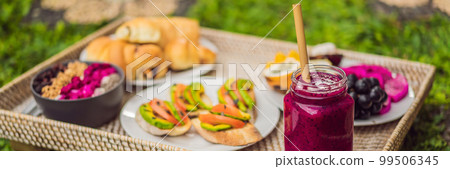 BANNER, LONG FORMAT Breakfast on a tray with fruit, buns, avocado sandwiches, smoothie bowl standing on the grass 99506345
