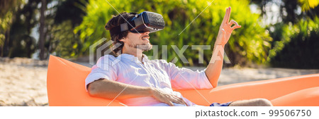 BANNER, LONG FORMAT Summer lifestyle portrait of man sitting on the orange inflatable sofa and uses virtual reality headset on the beach of tropical island. Relaxing and enjoying life on air bed BANNER, LONG FORMAT Summer lifestyle portrait of man sitting on the orange inflatable sofa and uses virtual reality headset on the beach of tropical island. Relaxing and enjoying life on air bed 99506750