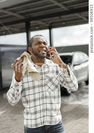 Man wiping luxury car on a car wash using a rag. Black man talking on the phone and holding in his hands rag for wiping his car. Man wearing plaid shirt and jeans. 99506915