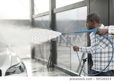 Man washing luxury car on a car wash with a pressure washer. Black man using high pressure hose with water. Man wearing plaid shirt. 99506924