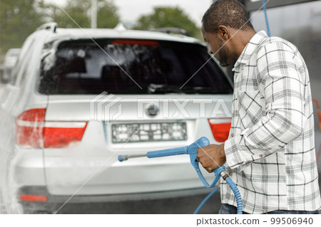 Man washing luxury car on a car wash with a pressure washer. Black man using high pressure hose with water. Man wearing plaid shirt. Man washing luxury car on a car wash with a pressure washer. Black man using high pressure hose with water. Man wearing plaid shirt. 99506940