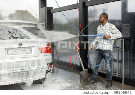 Man washing luxury car on a car wash with a pressure washer. Black man using high pressure hose with water. Man wearing plaid shirt. 99506941