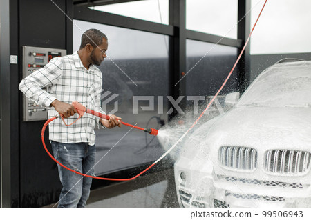 Man washing luxury car on a car wash with a pressure washer. Black man using high pressure hose with soap. Man wearing plaid shirt. Man washing luxury car on a car wash with a pressure washer. Black man using high pressure hose with soap. Man wearing plaid shirt. 99506943
