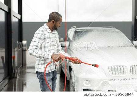 Man washing luxury car on a car wash with a pressure washer. Black man using high pressure hose with soap. Man wearing plaid shirt. Man washing luxury car on a car wash with a pressure washer. Black man using high pressure hose with soap. Man wearing plaid shirt. 99506944