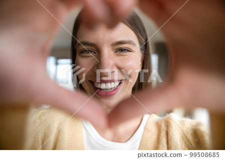 Cheerful beautiful young woman looking at camera through hand heart Cheerful beautiful young woman looking at camera through hand heart 99508685