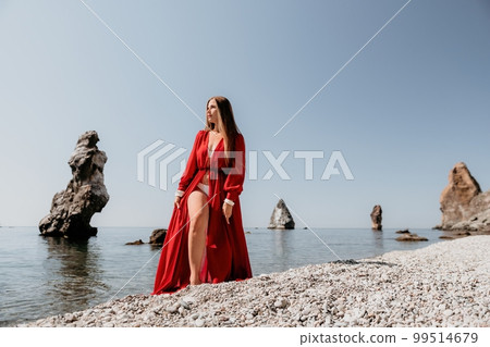 Woman travel sea. Happy tourist in red dress enjoy taking picture outdoors for memories. Woman traveler posing on the rock at sea bay surrounded by volcanic mountains, sharing travel adventure journey 99514679