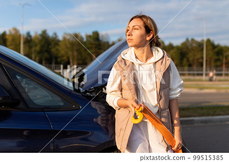 an adult Caucasian businesswoman with a cable for transporting a car stands at the raised hood of a an adult Caucasian businesswoman with a cable for transporting a car stands at the raised hood of a 99515385