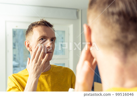Middle aged man apply face tonic with cotton pad, then massage with hands. Male portrait in mirror. Face cleansing. 99516046
