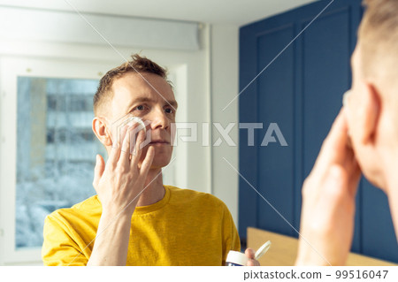 Mature man apply face tonic with cotton pad, then massage with hands. Male portrait in room mirror. Face cleansing. 99516047