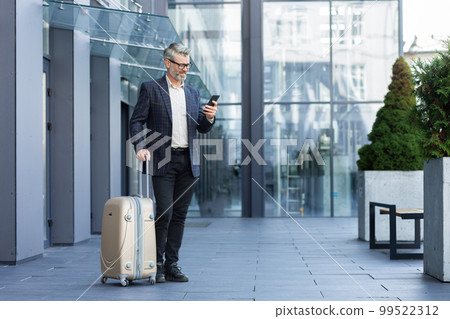 A man in a business suit with a large travel suitcase and a phone is typing a message and booking a taxi, a boss on a business trip, a mature businessman outside a large hotel. A man in a business suit with a large travel suitcase and a phone is typing a message and booking a taxi, a boss on a business trip, a mature businessman outside a large hotel. 99522312