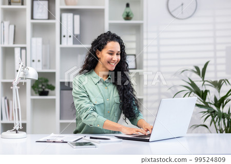 Young successful hispanic businesswoman working in bright home office indoors sitting at table with documents and reports, woman using laptop at work happy smiling. 99524809