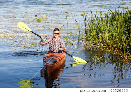 A young Caucasian man sits in a kayak and paddles. The concept of water entertainment. A young Caucasian man sits in a kayak and paddles. The concept of water entertainment. 99526923