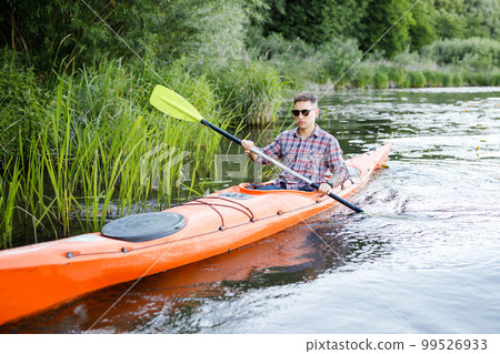 A young Caucasian man sits in a kayak and paddles. The concept of water entertainment. A young Caucasian man sits in a kayak and paddles. The concept of water entertainment. 99526933