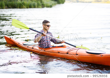 A young Caucasian man sits in a kayak and paddles. The concept of water entertainment. A young Caucasian man sits in a kayak and paddles. The concept of water entertainment. 99526938