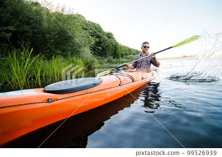 A young Caucasian man sits in a kayak and paddles. The concept of water entertainment. A young Caucasian man sits in a kayak and paddles. The concept of water entertainment. 99526939