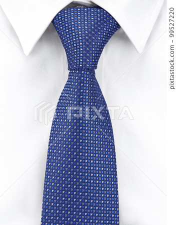 Close-up of a necktie made of blue silk with a geometric pattern, tied with a Windsor knot on a white shirt with a classic collar. Vertical image. 99527220