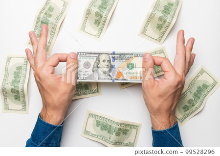 Top-down first-person photo of a hand holding a hundred dollar bill in front of a pattern of money 99528296