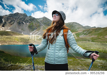 Portrait of a tourist with a backpack, front view. A young girl leads an active lifestyle, walks along a mountain path with trekking poles, mountain peaks and a lake in the background 99528298