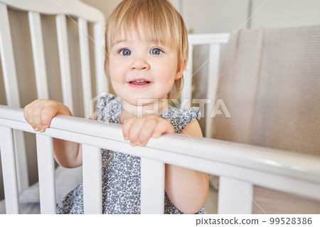 Happy baby girl one and half standing in the crib. Smiling 18 months child playing in sunny bedroom. Baby cot in the parent's bedroom 99528386