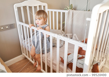 Happy baby girl one and half standing in the crib. Smiling 18 months child playing in sunny bedroom. Baby cot in the parent's bedroom 99528400