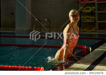 Resting. Young woman, professional female swimming athlete in cap and goggles posing in pool indoor. Concept of sport, endurance, competition Resting. Young woman, professional female swimming athlete in cap and goggles posing in pool indoor. Concept of sport, endurance, competition 99528876