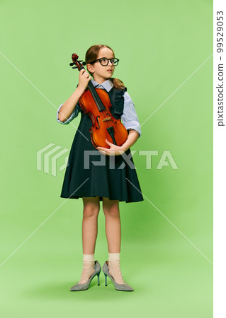 Cute little girl, talanted musician wearing huge mother's heels shoes playing violin, having fun isolated over green background. Education, music, classic art, new vision, fun concept 99529053