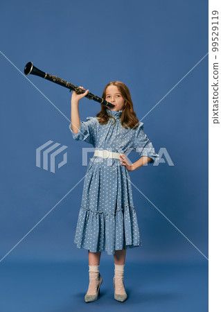 Monochrome portrait of charming nice little girl in dress and big shoes playing on clarinet over blue background. Music, fashion, hobby concept 99529119