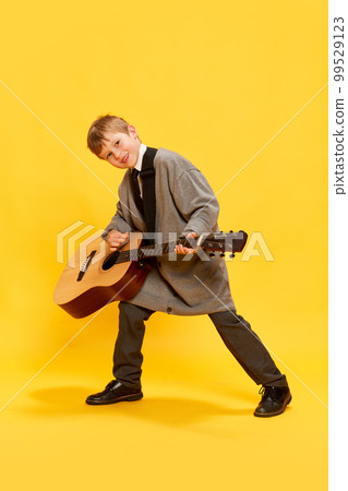 Charming little boy, funny musician wearing huge oversize clothes playing guitar, having fun isolated over yellow background. Pop art, music, new vision, fun concept 99529123