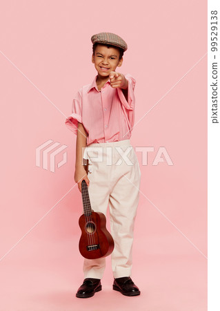 Happy emotional little african boy in retro style clothes and cap posing with ukulele guitar isolated on pink background. 99529138