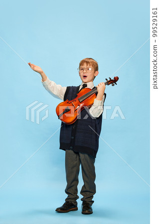 Happy kid, little musician wearing big size clothes posing with violin isolated over blue background. Education, music, classic art, new vision, fun concept 99529161
