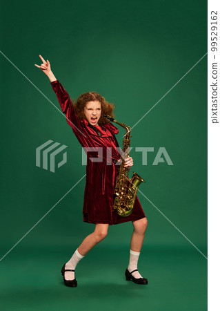 Curly redhaired happy school age girl wearing festive dress playing on saxophone over green background. Studying, fashion, art, music and creative style 99529162