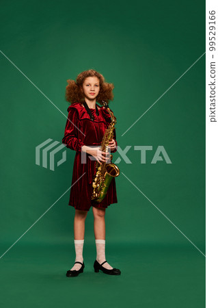 Curly redhaired happy school age girl wearing festive dress playing on saxophone over green background. Studying, fashion, art, music and creative style Curly redhaired happy school age girl wearing festive dress playing on saxophone over green background. Studying, fashion, art, music and creative style 99529166