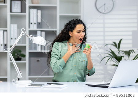 Woman with sore throat at workplace inside office, business woman with laptop working at work, Hispanic woman using medicine to relieve sore throat. Woman with sore throat at workplace inside office, business woman with laptop working at work, Hispanic woman using medicine to relieve sore throat. 99531808