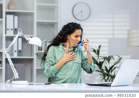 Asthma attack in Hispanic business woman inside office at workplace with laptop, woman uses inhaler with medication to ease breathing. Asthma attack in Hispanic business woman inside office at workplace with laptop, woman uses inhaler with medication to ease breathing. 99531810