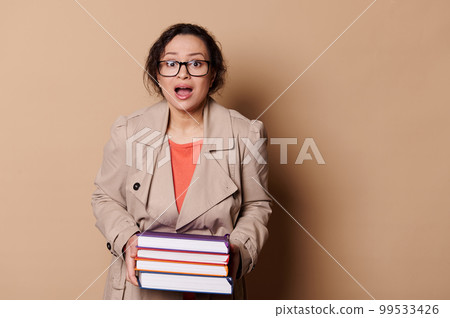 Astonished multi-ethnic woman wearing beige coat and eyeglasses, a school teacher holding heavy volumes of hardcover books, expressing stupefaction looking at camera, isolated over cream background Astonished multi-ethnic woman wearing beige coat and eyeglasses, a school teacher holding heavy volumes of hardcover books, expressing stupefaction looking at camera, isolated over cream background 99533426