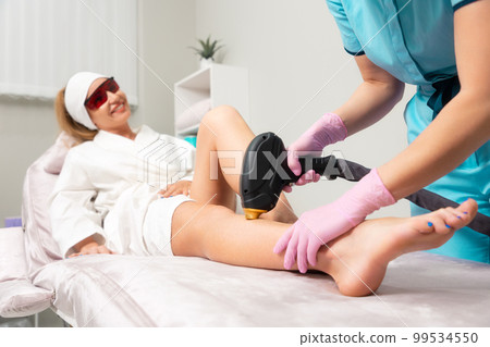 Cosmetologist in pink gloves removes hair with laser epilation to the client's leg. The concept of professional care procedure and cosmetology 99534550