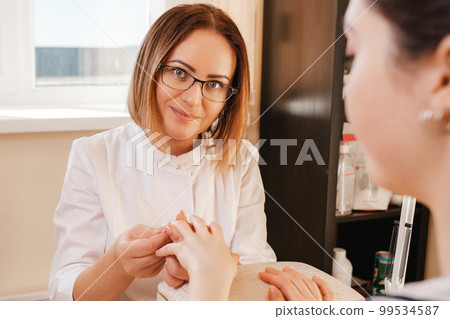 Medicine, cosmetology and manicure. A woman manicure in glasses, a mask and a white coat does a manicure to a client in the salon. View from over the visitor's shoulder. Close up Medicine, cosmetology and manicure. A woman manicure in glasses, a mask and a white coat does a manicure to a client in the salon. View from over the visitor's shoulder. Close up 99534587