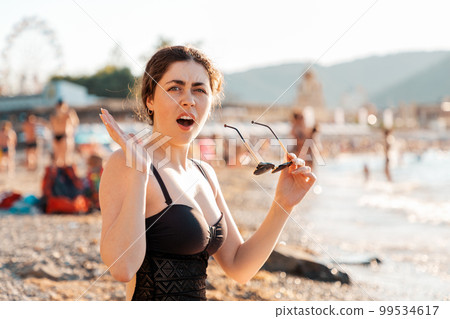 Human emotion. A woman is sitting on the beach in a swimsuit, dissatisfied with the spread of her hands, expressing protest and indignation 99534617