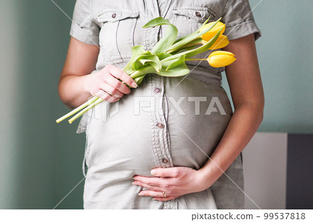 Pregnant woman with tulips flowers holding hands on belly on a pastel background. Mothers day or motherhood concept. Pregnant woman with tulips flowers holding hands on belly on a pastel background. Mothers day or motherhood concept. 99537818