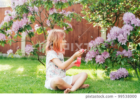 Cute little girl with toned red hair blowing soap bubbles while sitting on a green lawn in the garden. Having fun during school holidays at home. Offline activity for kids. Happy childhood concept 99538000