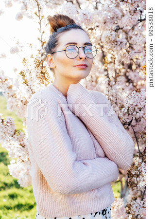 Portrait of young caucasian brunette hipster girl in glasses and light pink clothes near blooming tree with sunset light and sunbeams. Generation Z girl enjoy spring mood. Vertical card. 99538018