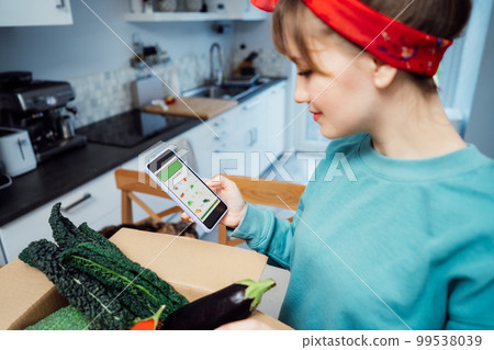 Online home food delivery. Focus on active app with order list on the phone screen. Woman checking her order. Cardboard box with fresh vegetables standing on the kitchen table. Local farmer food. Online home food delivery. Focus on active app with order list on the phone screen. Woman checking her order. Cardboard box with fresh vegetables standing on the kitchen table. Local farmer food. 99538039