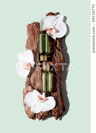 Two green glass cosmetic bottles on textured tree bark podium with white orchid flowers on pastel mint background. Demonstration of cosmetic products with copy space in flat lay style. 99538770