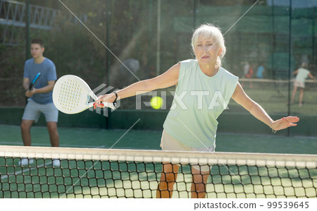 Sporty mature woman padel player hitting ball with a racket on hard court 99539645