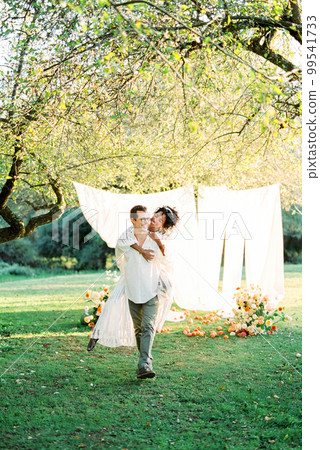 Man carries a laughing woman on his back against the background of white curtains in a green garden Man carries a laughing woman on his back against the background of white curtains in a green garden 99541733