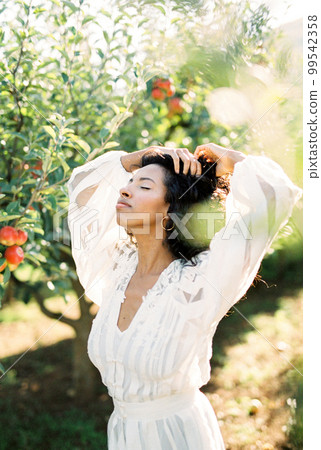 Woman in a white dress touches her hair with her hand while standing in an apple orchard 99542358