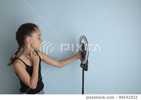 Little emotional teenage girl in a black t-shirt on an isolated blue background. Studio portrait of a young smiling teenage girl, vlogger, with a ring flash lamp and a smartphone. Girl takes a selfie. Little emotional teenage girl in a black t-shirt on an isolated blue background. Studio portrait of a young smiling teenage girl, vlogger, with a ring flash lamp and a smartphone. Girl takes a selfie. 99542832