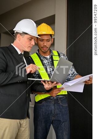 Determined Asian male foreman looking at laptop screen, working with a Caucasian businessman Determined Asian male foreman looking at laptop screen, working with a Caucasian businessman 99547209